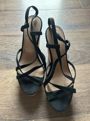 Svarta kilklackade sandaler - Snygga svarta sandaler med korsade remmar i mockaimitation och hög kilklack i korkmaterial. Öppen tå och justerbar rem runt ankeln ger en trendig och bekväm look. Perfekta för sommarens alla outfits.