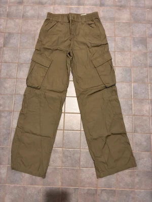 Olivgröna cargobyxor med fickor - Säljer ett par olivgröna cargobyxor med flera stora fickor på sidorna och resår i midjan. Byxorna har en avslappnad passform och är perfekta för en streetwear-look. Materialet känns lätt och bekvämt, perfekt för dig som gillar praktiska och snygga byxor.