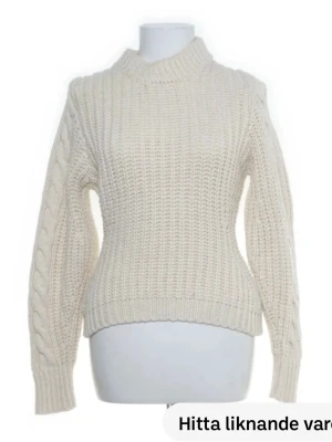 Stickad tröja - Säljer en krämvit stickad tröja med hög hals och snygga flätade detaljer längs ärmarna. Tröjan har ribbade muddar vid ärmslut och nederkant, och är gjord i ett tjockare garn som ger en mysig känsla. Perfekt för kyliga dagar.