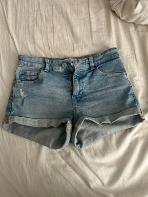 Blå denimshorts från Pull&Bear - Säljer ett par klassiska blå denimshorts från Pull&Bear med medellåg midja och uppvikta benslut. Shortsen har fem fickor, bälteshällor och en liten sliten detalj framtill. Perfekta för sommaren och lätta att matcha med olika stilar.