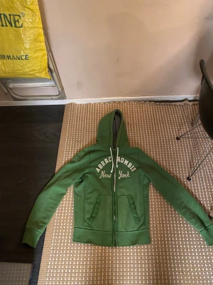 Grön hoodie från Abercrombie - Snygg grön hoodie från Abercrombie med dragkedja och vit broderad text 'Abercrombie New York' på bröstet. Utrustad med huva och vita snören samt två fickor framtill. Perfekt för en avslappnad och cool stil.