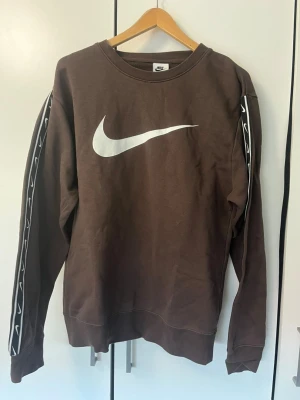 Brun Nike sweatshirt med logga - Brun sweatshirt från Nike med stor vit Swoosh-logga på bröstet och svarta band med vita Swoosh-detaljer längs ärmarna. Klassisk rund halsringning och ribbade muddar. Perfekt för en sportig och avslappnad stil.