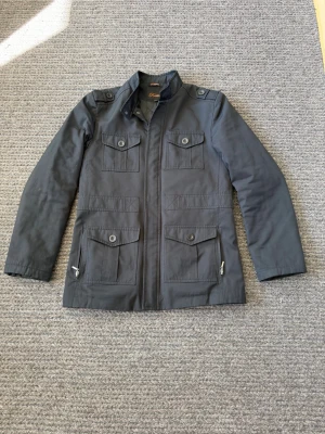 Overshirt - Snygg mörkgrå vindjacka med fyra stora fickor framtill, knappar och dragkedja. Jackan har hög krage och axelklaffar för en cool look. Tillverkad i ett slitstarkt material som passar perfekt till vardagsoutfits.