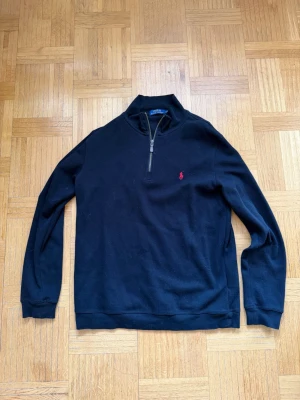 Svart half-zip tröja från Polo Ralph Lauren - Svart långärmad tröja från Polo Ralph Lauren med half-zip och röd broderad logga på bröstet. Klassisk rak passform och ribbade muddar. Perfekt för en clean och stilren look. Använd med vård, storlek L. Skriv vid funderingar ☺️