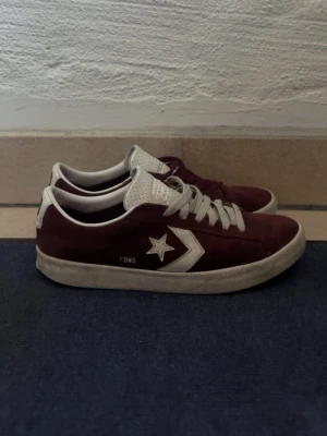 Bordeaux Converse CONS sneakers - Snygga bordeauxfärgade Converse CONS sneakers med vit sula och vita detaljer. Skorna har klassisk låg profil, vit stjärnlogga på sidan och olika färg på skosnörena – ett vitt och ett bordeaux. Ovandelen är i mocka och tungan är perforerad för extra ventilation.