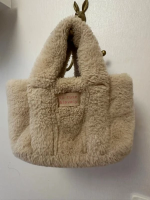 Beige fluffig handväska från Zaxy - Supersöt beige handväska i mjuk fuskpäls från Zaxy. Väskan har två handtag, magnetknäppning och en etikett med texten 'so cute & lovely' på framsidan. Perfekt för dig som gillar gulliga accessoarer och vill ha något mysigt att bära dina saker i.