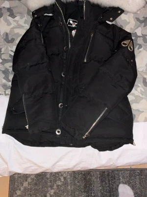 Moose Knuckles jacka - Säljer en svart dunjacka från Moose Knuckles med avtagbar huva och fuskpäls, flera dragkedjeförsedda fickor och snygga metalliska detaljer på ärmen. Jackan har en klassisk puffer-modell och är perfekt för kalla vinterdagar.