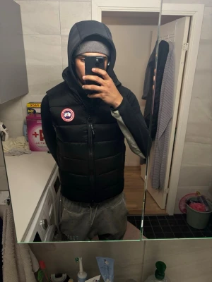 Svart dunväst från Canada Goose - Säljer en svart dunväst från Canada Goose med klassisk logotyp på bröstet. Västen har en rak passform, dragkedja framtill och är tillverkad i ett slitstarkt material med dunfyllning som håller värmen. Perfekt för lager-på-lager och streetwear-stil.