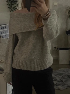 Ljusgrå off shoulder stickad tröja - Mysig ljusgrå stickad tröja med bred off shoulder-krage och långa ärmar. Tröjan har en avslappnad passform och ribbstickade muddar vid ärmslut och nederkant. Perfekt för en trendig och bekväm look.