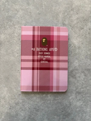 A Bathing Ape 2009 Summer Style Manual - Säljer en unik stilmanual från A Bathing Ape, 2009 Summer Edition. Boken har ett rutigt omslag i olika nyanser av rosa och rött med rundade hörn. Perfekt för dig som gillar streetwear och vill ha inspiration från BAPE.