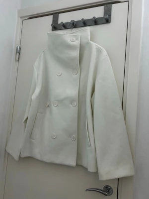 Vit skepparkavaj från BUBBLEROOM, strl 42 - Stilren vit skepparkavaj från BUBBLEROOM med dubbelknäppning och bred krage som kan bäras uppvikt eller nedvikt. Jackan har raka ärmar, två sidofickor och är tillverkad i ett mjukt material. Perfekt för dig som gillar minimalistisk och modern design. Nypris är ca 1000kr 