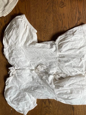 Vit offshoulder blus med puffärm - Söt vit offshoulder blus med puffiga korta ärmar och knytning framtill. Tillverkad i ett lätt och luftigt tyg, perfekt för en trendig och avslappnad look. Passar till både jeans och kjol för en cool stil.