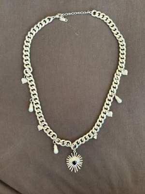 Chunky halsband med hjärthänge - Snyggt chunky halsband i silverfärgad metall från Wos. Flera små berlocker i form av hjärtan och pärlor, en pärla har ramlat av.  Mittendelen har ett solformat hänge med svart hjärta och. Skicket är mycket bra och endast använt ett få tal gånger utöver pärlan som trillat av. 