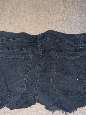 Svarta jeansshorts från Ashley Max - Säljer ett par svarta jeansshorts från Ashley Max med rå kant nertill och klassisk femficksdesign. Shortsen har bälteshällor, knapp och dragkedja framtill. Perfekta för en avslappnad och cool stil.