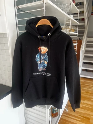 Svart Ralph Lauren hoodie - Svart hoodie från Ralph Lauren med den ikoniska Polo Bear-trycket på bröstet. Hoodien har huva med dragsko, stor magficka och ribbade muddar. Perfekt för dig som gillar streetwear och vill ha en klassisk look med en twist.