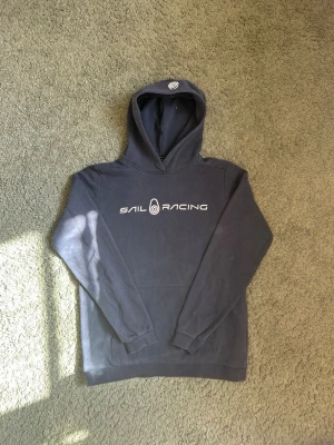 Mörkblå hoodie från Sail Racing - Snygg mörkblå hoodie från Sail Racing med vit logga tryckt på bröstet. Klassisk huva och känguruficka framtill. Tillverkad i mjuk bomull och polyester blandning som känns skön mot huden. Perfekt för en avslappnad och sportig stil.