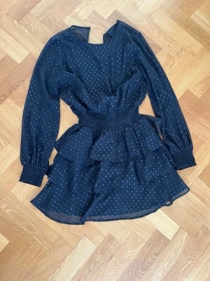 Svart volangklänning med prickar - Söt svart kortklänning med små prickar och volangdetaljer. Klänningen har långärmade ärmar med resår vid handlederna och markerad midja med smock. Tillverkad i ett lätt, transparent material med underklänning.