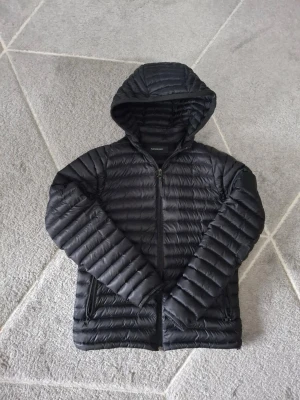 Svart dunjacka med huva från Peak Performance - Snygg svart dunjacka från Peak Performance med quiltad design och huva. Jackan har dragkedja framtill, två sidofickor med dragkedja och diskret logga på ärmen. Perfekt för kalla dagar och enkel att matcha med olika outfits.