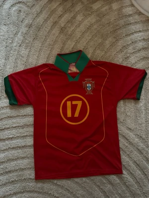 Portugal fotbollströja C. Ronaldo 17 - Säljer en röd Portugal fotbollströja med nummer 17 och C. Ronaldo på ryggen. Tröjan har gröna detaljer på krage och ärmslut samt Portugals landslagsemblem på bröstet. Tillverkad i lätt och ventilerande syntetmaterial, perfekt för träning eller match.
