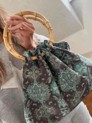 Mönstrad handväska med bambuhandtag - Unik handväska med rundade bambuhandtag och tyg i brunt med turkost paisleymönster. Väskan har en mjuk, påsig form och dragskoöppning. Perfekt accessoar för dig som gillar bohemisk och retro stil.