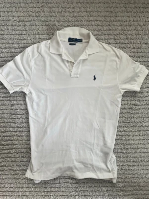 Vit pikétröja från Polo Ralph Lauren - Klassisk vit pikétröja från Polo Ralph Lauren med blå logga broderad på bröstet. Tröjan har krage, korta ärmar och knappar vid halsen. Classic fit-modell i mjuk bomull som passar perfekt till jeans eller shorts. PRIS KAN DISKUTERAS