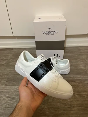 Valentino skor - Snygga vita sneakers från Valentino Garavani med en bred svart rem över vristen dekorerad med nitar. Skorna har snörning och är tillverkade i skinn för en lyxig känsla. Perfekta för dig som vill ha en clean men ändå edgy stil.