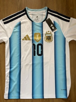 Argentina Messi Landslagströja - Säljer en Argentina matchtröja från Adidas med Messi och nummer 10 på ryggen. Tröjan har blå och vita vertikala ränder, svarta detaljer och guldiga loggor på bröstet. Tillverkad i lätt och ventilerande material med Climalite-teknologi.      Tröjan är i storlek S och är aldrig använd. Perfekt inför VM2026!                                                                   Skriv om ni har några funderingar!😃