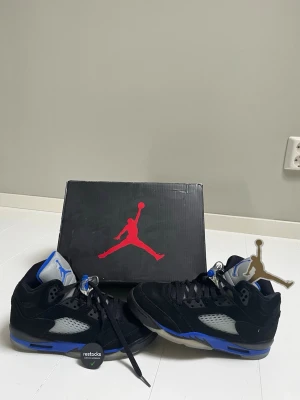 Air Jordan 5 Racer Blue - Snygga Air Jordan 5 Retro Racer Blue sneakers i svart mocka med blå och grå detaljer. Skorna har en ikonisk siluett, genomskinliga meshpaneler på sidorna och reflekterande plös med Jumpman-logga. Sulan är svart och blå med isiga detaljer. Perfekta för dig som gillar streetwear. Storlek 38,5 EU