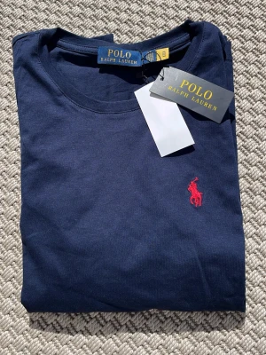Marinblå t-shirt från Polo Ralph Lauren - Snygg marinblå t-shirt från Polo Ralph Lauren med klassisk röd broderad logga på bröstet. Tillverkad i mjuk bomull och har en normal passform med korta ärmar. Perfekt basplagg för en clean och stilren look.