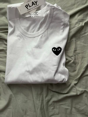 Vit Comme des Garçons PLAY t-shirt - Säljer en vit t-shirt från Comme des Garçons PLAY med det ikoniska svarta hjärta-trycket på bröstet. Klassisk rund halsringning och korta ärmar. Tillverkad i mjuk bomull för skön känsla och stilren look.