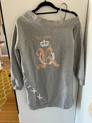 Grå loungewear-klänning med tryck - Mysig grå loungewear-klänning med stort R-tryck, krona och texten EST. 1988 framtill. Off-shoulder design och dekorativa vita banddetaljer nertill. Perfekt för avslappnade dagar hemma och en kall sommarkväll. Helt oanvänd med prislapp kvar.