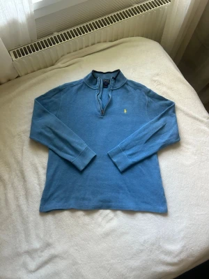 Blå halvzip tröja från Polo Ralph Lauren - Snygg blå långärmad tröja från Polo Ralph Lauren med halv dragkedja och klassisk gul logga på bröstet. Tröjan har ribbade muddar och står upp lite i kragen. Perfekt för en chill och stilren look.