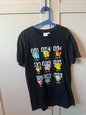 Svart Pokémon t-shirt med tryck - Svart t-shirt från Pokémon med färgglatt tryck av flera klassiska Pokémon och deras nummer på framsidan. På baksidan står det 'POKÉMON' i stora bokstäver. T-shirten har rund hals och korta ärmar, tillverkad i mjuk bomull.