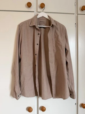 Beige flanellskjorta från Jack & Jones - Säljer en beige långärmad flanellskjorta från Jack & Jones Essentials. Skjortan är i mjuk 100% bomull med klassisk krage, bröstficka och knappar framtill. Perfekt för dig som gillar en enkel och stilren look.