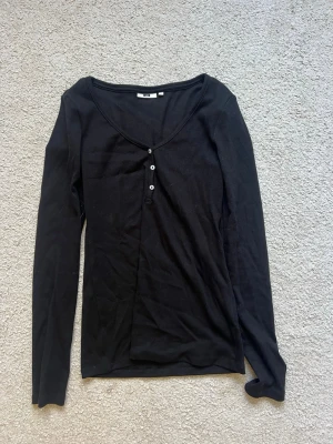 Svart långärmad topp med knappar - En svart långärmad topp med djup v-ringning och tre små knappar framtill. Tunn och mjuk i materialet, perfekt för lager på lager eller som den är. Enkel och stilren design som passar till det mesta.