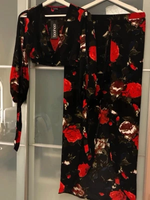 Lång blommig kjol från Boohoo - Säljer ett snyggt tvådelat set från Boohoo med svart botten och stora röda och vita rosor. Setet består av en långärmad crop top med omlott-ringning och en matchande lång skort ( shorts som likar kjol) . Materialet är lätt och flowy, perfekt för att sticka ut med ett färgstarkt mönster.