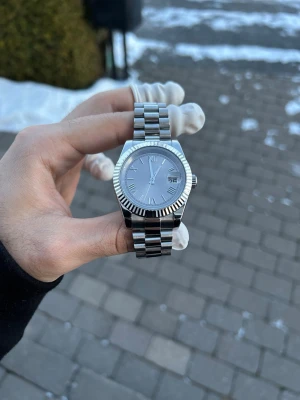 Silverfärgad klocka med datumfönster - Stilren klocka i silver med klassisk länkarmband och räfflad bezel. Urtavlan är också silverfärgad med romerska siffror och datumfönster vid klockan tre. Perfekt accessoar för dig som gillar en clean och tidlös look.