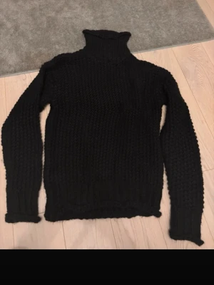 Svart chunky stickad polotröja - Mysig svart stickad polotröja med grovstickad struktur och hög, ribbad krage. Tröjan har långa ärmar och ribbade muddar vid ärmslut och nederkant. Perfekt för dig som gillar en oversized och varm look. Materialet ser ut att vara ull eller ullmix.