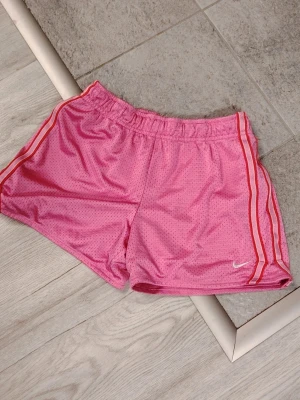Rosa sportiga shorts från Nike💕  - Snygga rosa meshshorts från Nike med vita och röda ränder längs sidorna. Shortsen har elastisk midja och är tillverkade i ett lätt och ventilerande syntetmaterial, perfekt för träning eller sportiga outfits😎🩷 Klassisk Nike-logga broderad på benet. Kolla på också andra ads! ☀️  Du får -10% rea via bundle💞 