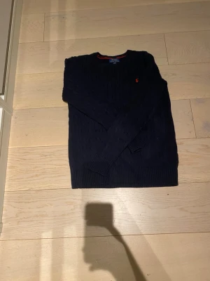  HELT NY! Mörkblå kabelstickad tröja Polo Ralph Lauren  - Snygg mörkblå kabelstickad tröja från Polo Ralph Lauren med klassisk röd logga broderad på bröstet. Tröjan har rund halsringning, långa ärmar och ribbade muddar. Perfekt för dig som gillar stilrena och tidlösa plagg.