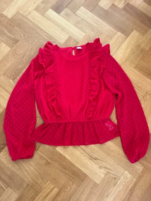 Röd volangblus med prickigt mönster - Supersöt röd blus med volanger längs axlarna och ett diskret prickigt mönster. Blusen har resår i midjan för en snygg siluett och långa puffiga ärmar med resår vid ärmslut. Materialet är lätt och ger en luftig känsla. Perfekt för dig som vill sticka ut lite extra. I storlek 170 men passar som en XS-S.