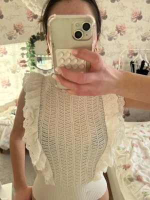 Vit ärmlös topp med spetsvolang - Supersöt vit topp med breda spetsvolanger längs axlarna och ett hålmönstrat tyg. Toppen är ärmlös och har en ribbad kant nedtill. Perfekt för dig som gillar romantisk och feminin stil.