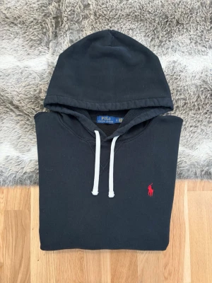 Svart Hoodie Från Polo Ralph Lauren - Hej! 👋 Säljer en klassisk svart Polo Ralph Lauren hoodie i storlek L. Den har den ikoniska röda pony-loggan broderad på bröstet 🐎 Tröjan är i gott skick, djupsvart i färgen och med den mjuka fleece-insidan intakt ✨ Sitter oversized på mig (Modell: 177cm), så den passar perfekt för en mer avslappnad look eller för dig som drar Large 👕 Ingen QR-kod på denna modell, men alla tvättlappar sitter kvar och garanterar att den är 100% äkta ✅ Fraktar snabbt! 🚛📦💬💯 