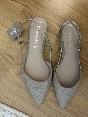 Silvriga/rosa ballerinaskor från Tamaris - Snygga ballerinaskor från Tamaris i glittrigt silvertyg (rosa också)  med spetsig tå och smala remmar runt hälen. Skorna har en elegant och modern look! 