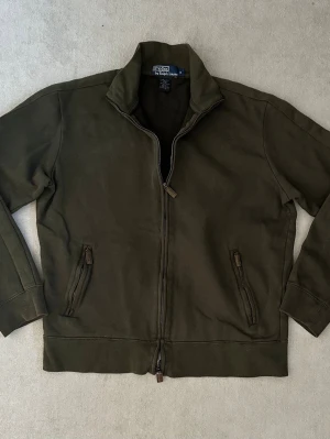 Mörkgrön ziptröja från Polo Ralph Lauren - Säljer en mörkgrön ziptröja från Polo Ralph Lauren med hög krage och två fickor med dragkedja. Tröjan har en enkel och stilren design, perfekt för lager på lager. Tillverkad i mjukt bomullsmaterial och har ribbade muddar vid ärmslut och nederkant. Tröjan är i 9/10 i skick. Är i storlek M men passar även S!