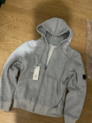 Grå hoodie från C.P. Company - Snygg grå hoodie från C.P. Company med dragkedja framtill, huva med dragsko och ribbade muddar. Klassisk patch med logga på ärmen och två fickor fram. Tillverkad i mjuk bomullsblandning för skön känsla.
