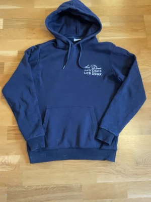 Mörkblå hoodie från Les Deux - Mörkblå hoodie från Les Deux med vit logga tryckt på bröstet. Klassisk känguruficka framtill, ribbade muddar och justerbar dragsko i huvan. Tillverkad i mjukt bomullsmaterial för en skön och avslappnad look.