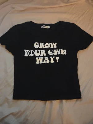 Svart kort t-shirt med tryck - Svart t-shirt med vit text 'Grow Your Own Way!' och symboler på bröstet. Klassisk rund halsringning och korta ärmar. Perfekt för dig som gillar retrostil och statement-plagg. 