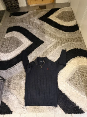 Svart kabelstickad tröja Polo Ralph Lauren - Kabelstickad svart tröja från Polo Ralph Lauren med halv dragkedja och röd broderad logga på bröstet. Tröjan har lång ärm, ribbade muddar och klassisk passform. Perfekt för dig som gillar stilrena och tidlösa plagg.