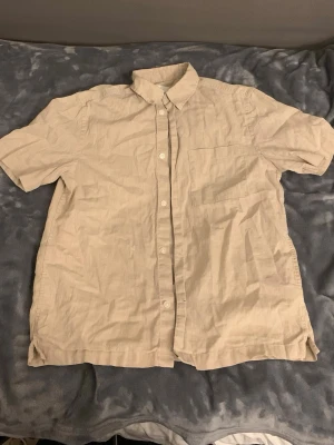 Beige kortärmad linneskjorta - Snygg beige skjorta i linne med korta ärmar och klassisk krage. Skjortan har knappar framtill och en bröstficka på vänster sida. Perfekt för varma dagar och enkel att styla till olika outfits.